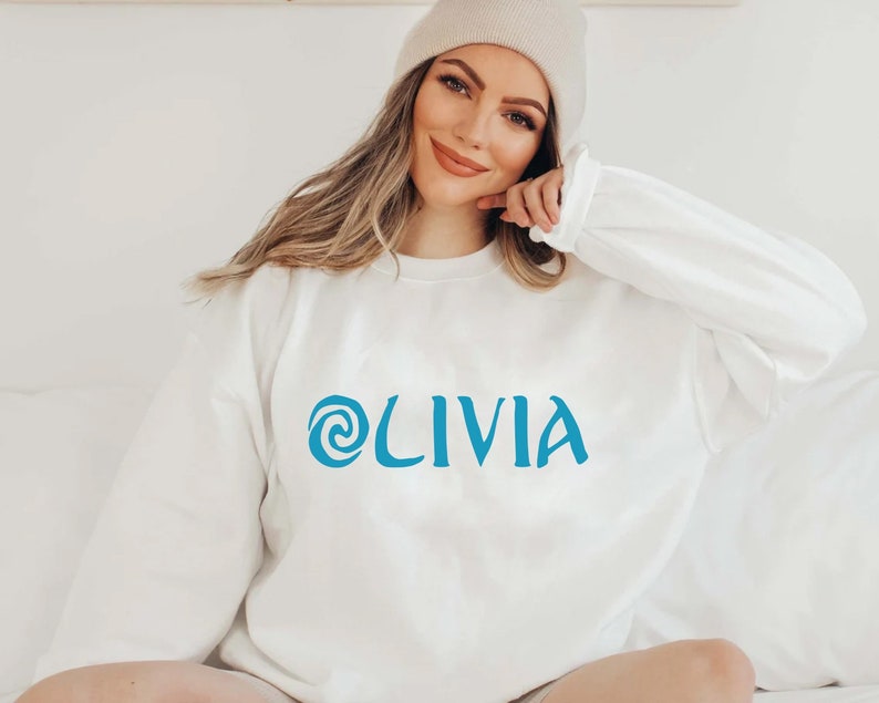 May include: A white sweatshirt with a blue swirl design and the name 'OLIVIA' printed on the front.