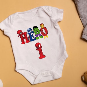 May include: White baby onesie with a graphic of the word "HERO" spelled out with different superhero characters. The number "1" is also spelled out with a Spiderman character.