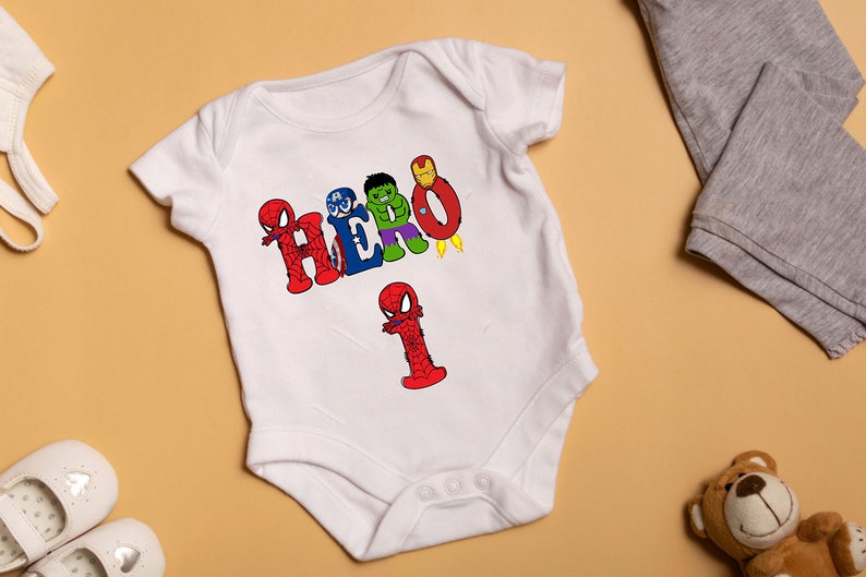 May include: White baby onesie with a graphic of the word "HERO" spelled out with different superhero characters. The number "1" is also spelled out with a Spiderman character.