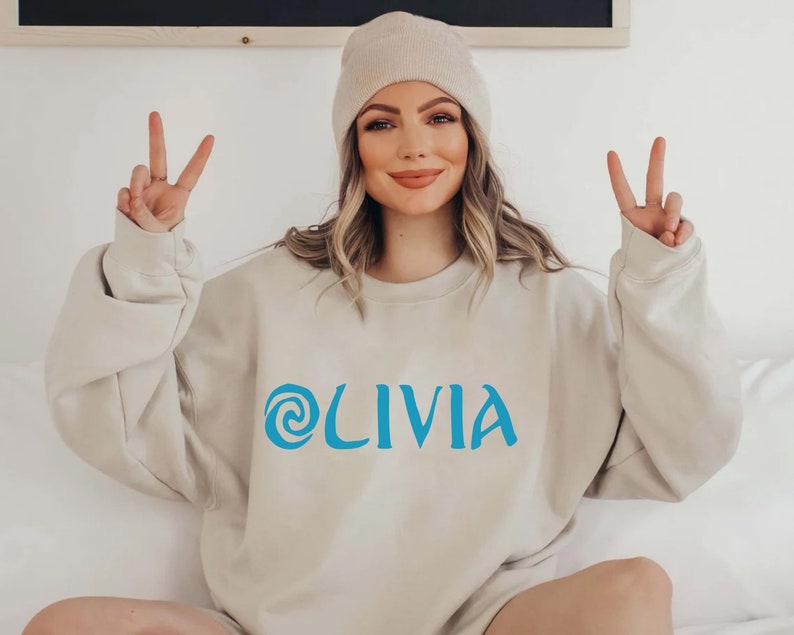 May include: A light beige sweatshirt with a blue swirl design and the word 'OLIVIA' printed on the front.