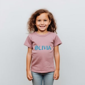 May include: A girl wearing a mauve t-shirt with a blue swirl and the word 'OLIVIA' printed on the front. The girl is smiling and looking at the camera.