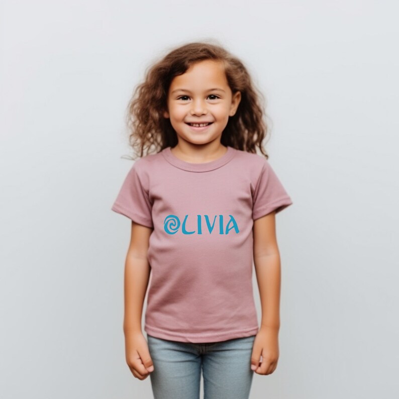May include: A girl wearing a mauve t-shirt with a blue swirl and the word 'OLIVIA' printed on the front. The girl is smiling and looking at the camera.