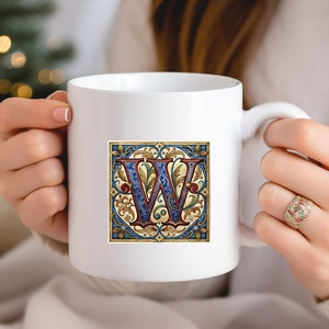 May include: White ceramic mug with a colorful illustration of the letter "W" in a decorative frame. The letter is blue and red with gold accents. The frame is blue, gold, and green.