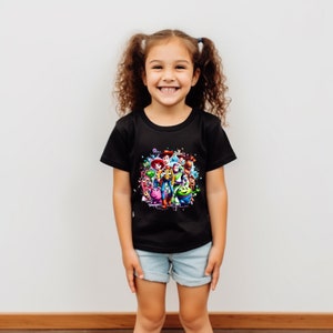 Pu&ograve; includere: Un bambino indossa una maglietta nera con una grafica colorata di personaggi del franchise Toy Story. La grafica include Woody, Buzz Lightyear, Jessie e altri personaggi.