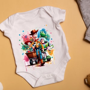Pu&ograve; includere: Body bianco per neonato con un design ad acquerello colorato che presenta Woody, Buzz Lightyear e Jessie del franchise Toy Story. I personaggi sono raffigurati in una posa giocosa, con Woody che tiene un carrello e Buzz Lightyear che vola.