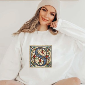 May include: White sweatshirt with a vintage-style illustration of a blue and gold letter 'S' surrounded by red and green floral details.