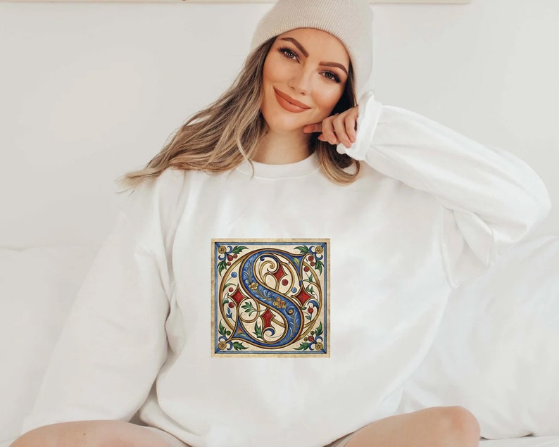 May include: White sweatshirt with a vintage-style illustration of a blue and gold letter 'S' surrounded by red and green floral details.