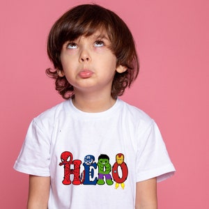 May include: A young child wearing a white t-shirt with a colorful graphic of superheroes spelling out the word "HERO".