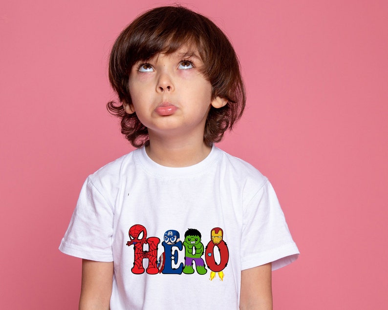 May include: A young child wearing a white t-shirt with a colorful graphic of superheroes spelling out the word "HERO".
