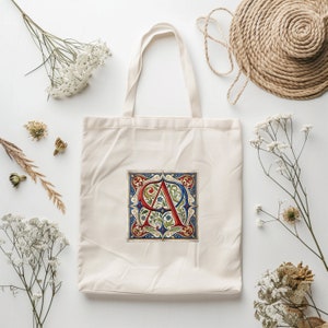 May include: A white canvas tote bag with a red, blue, and green floral design featuring the letter "A" in a decorative font. The bag has a long handle.