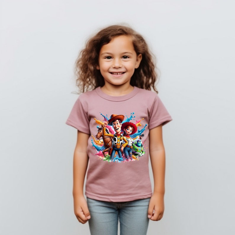 May include: A mauve t-shirt with a colorful graphic print of Woody, Jessie, and Buzz Lightyear from the Toy Story franchise. The characters are surrounded by colorful paint splatters.