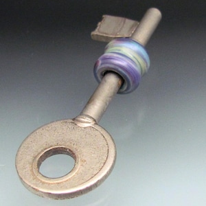 May include: A silver-toned key pendant with a decorative, multi-colored glass bead. The bead features swirls of blue, purple, and green. The key has a circular head with a hole and a rectangular top.