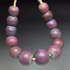 May include: A strand of handcrafted beads in shades of purple and blue. The round beads have a textured surface and are strung on a natural fiber cord. The beads are a mix of deep purple and lighter blue hues, creating a unique and artistic look.