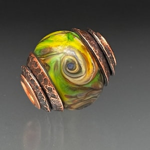 May include: A handcrafted glass bead with a swirling pattern of yellow, green, and brown colors. The bead is encircled by textured copper rings, creating a unique and artistic design. The bead has a small blue dot in the center.