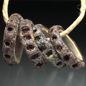 May include: Close-up of a set of five textured, dark-colored rings with irregular holes, strung on a natural-colored cord. The rings have a mottled appearance with hints of brown, purple, and blue, suggesting a handcrafted or aged aesthetic.