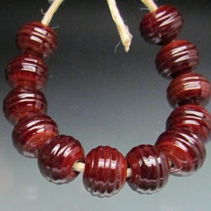 May include: A strand of deep red, ribbed glass beads. The beads are round with a grooved texture, strung together on a natural-colored cord. The beads have a glossy finish, reflecting light. The overall theme is jewelry making or crafting.