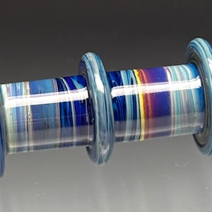 May include: A handcrafted glass plug with a cylindrical shape. The plug features a blue and white striped pattern with accents of yellow, orange, and purple. The ends and center ring are a solid light blue color.
