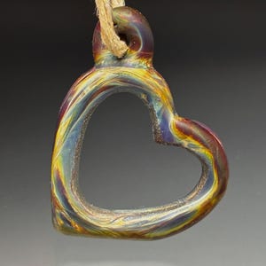 May include: A heart-shaped pendant made of iridescent glass, suspended by a natural fiber cord. The glass displays a swirling pattern of colors including yellow, red, blue, and purple. The pendant is a unique piece of jewelry.