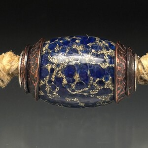 May include: A close-up of a necklace featuring three blue and beige speckled beads. The central bead is oval-shaped, flanked by two round beads. Each bead is accented with copper-colored metal rings and strung on a natural fiber cord.