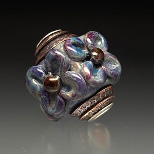 May include: A handcrafted glass bead with a floral design. The bead features swirling shades of purple, blue, and clear glass, accented with brown and copper-colored metal bands. A small, dark brown glass bead is at the center.