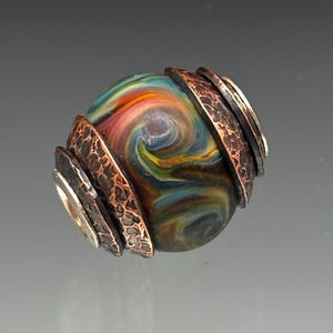 May include: A handcrafted glass bead with swirling colors of blue, yellow, orange, and green. The bead is accented with textured copper and silver metal rings, creating a unique and artistic design.