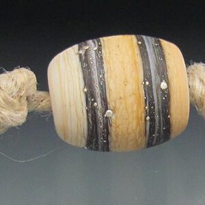 May include: A strand of handmade beads with a natural, rustic aesthetic. The beads are a mix of cylindrical and barrel shapes, featuring a cream-colored base with dark brown and gray stripes. The beads are strung on a natural fiber cord.