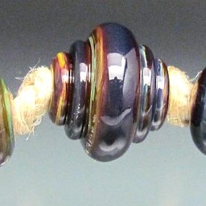 May include: A strand of colorful glass beads with a central, ridged bead. The beads are various colors, including brown, blue, and yellow, strung on a natural-colored cord. The beads have a glossy finish and reflect light.