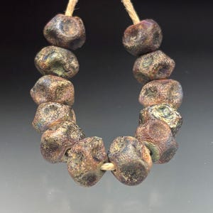 May include: A strand of dark, textured beads with a mottled appearance. The beads are primarily brown with hints of green and purple, strung on a natural-colored cord. The beads have an irregular, organic shape.