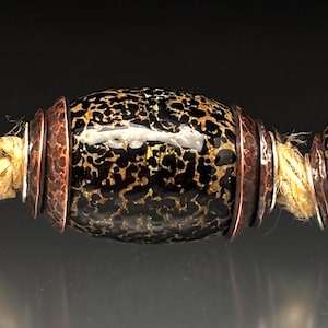 May include: Close-up of a beaded necklace featuring three dark, speckled beads. The central bead is oval-shaped, flanked by two round beads. Copper-colored metal rings separate the beads, strung on a natural-colored cord.