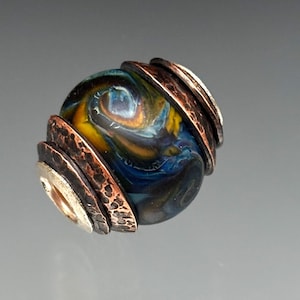 May include: A handcrafted glass bead with swirling blue, yellow, and orange colors. The bead is accented with textured copper and silver metal rings, creating a unique and artistic design.