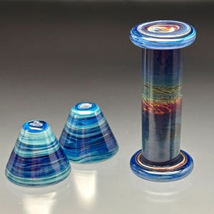 May include: Three pieces of handcrafted glass art. Two are cone-shaped, featuring blue and white swirling patterns. The third is a cylindrical piece with a blue top and base, and a central section with a mix of colors, including orange and green.
