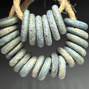 May include: A circular arrangement of blue-green beads with a textured surface, strung on a natural-colored rope. The beads are cylindrical and have a weathered appearance, suggesting an antique or handcrafted origin. The background is dark.