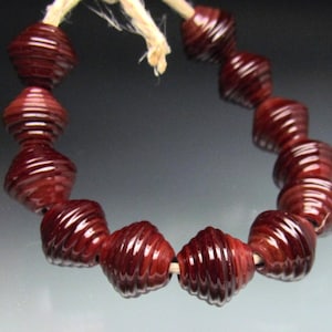 May include: A strand of dark red, spiral-shaped beads. The beads are strung together with a natural-colored cord. The beads have a glossy finish and are evenly spaced along the cord. The background is a gradient of gray and white.