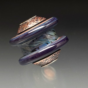 May include: A handcrafted glass bead with a swirling blue and black pattern, framed by two copper-colored metal rings. The bead has a dark purple, glossy finish and is designed with a unique, artistic shape.