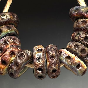 May include: A strand of dark brown, oval-shaped beads with three holes each. The beads have a textured surface and are strung on a natural-colored cord. The beads have a mix of colors, including brown, gold, and purple.