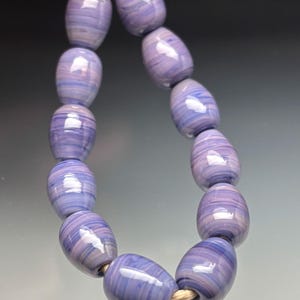 May include: A strand of lavender-colored, oval-shaped beads with a swirling pattern of light and dark purple and white. The beads are strung on a natural-colored cord, creating a necklace or bracelet.