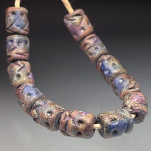 May include: A strand of handmade beads with a rustic, aged appearance. The beads are cylindrical, with a textured, weathered surface in shades of brown, blue, and purple. Each bead features carved details and is strung on a natural-colored cord. The beads are approximately 0.75 inches in length.