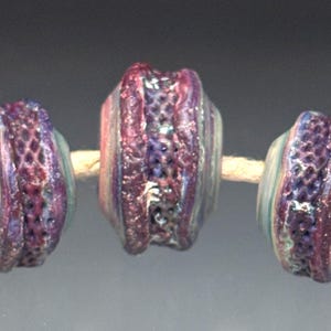 May include: Three handmade glass beads strung on a natural fiber cord. The beads feature a mix of purple, pink, and teal colors with a textured, honeycomb-like pattern around the center. The beads are cylindrical with rounded edges.