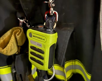 Puede incluir: Un micrófono de altavoz remoto Motorola XE500 de color amarillo neón brillante sujeto a una chaqueta de bombero negra con un clip giratorio de metal plateado. El dispositivo cuenta con una rejilla de altavoz negra, un mando y un cable.