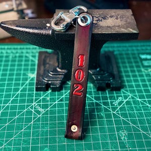 May include: A red and black leather keychain with the number "102" embossed in red. The keychain is attached to a silver metal key ring.