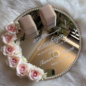 Puede incluir: Una bandeja redonda y espejada con borde dorado y ribete de perlas, adornada con rosas rosas. Muestra dos cajas de anillos beige y caligrafía blanca, incluyendo los nombres "Tamara Ahmad" y la fecha "24 de mayo de 2026".