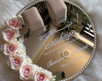 May include: A round, mirrored tray with a gold rim and pearl border, decorated with pink roses. The tray holds two beige ring boxes and features white calligraphy and the names "Tamara Ahmad" with the date "May 24, 2026".