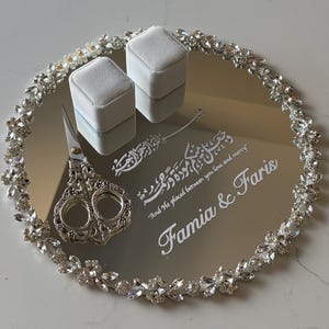Puede incluir: Una bandeja circular de espejo con un borde decorativo de cristales transparentes y detalles florales. Dos cajas de terciopelo gris claro para anillos y unas tijeras plateadas ornamentadas descansan sobre ella. El texto blanco dice Famia & Faris.