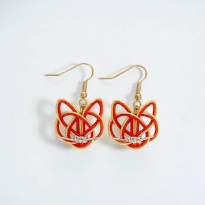 以下が含まれることがあります： 赤と金のデザインが施された、金色のドロップピアス。ピアスは、複雑な結び目と銀色のアクセントを持つ、様式化されたキツネの顔の形をしています。金色のフックピアスからぶら下がっています。