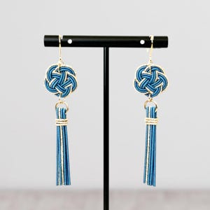May include: A pair of blue and gold earrings with a tassel. The earrings feature a knot design and are hanging from a black stand.