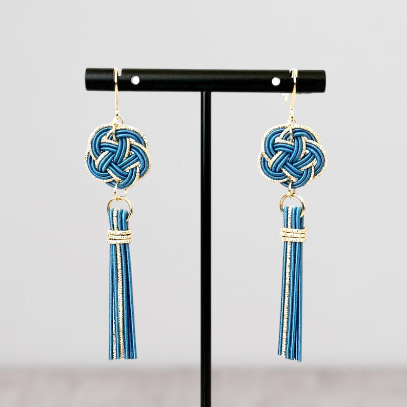 May include: A pair of blue and gold earrings with a tassel. The earrings feature a knot design and are hanging from a black stand.