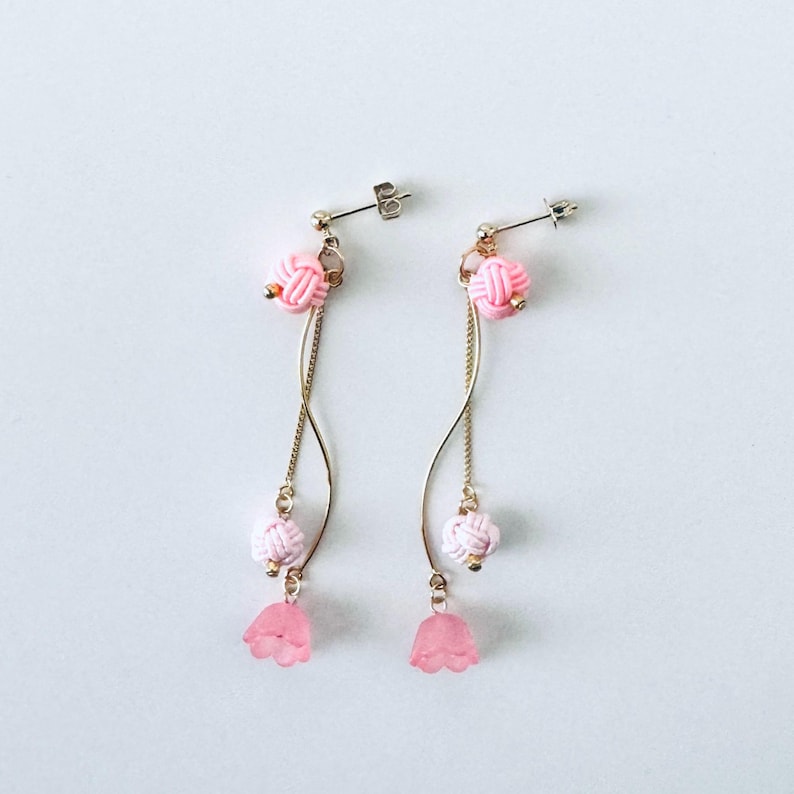 May include: A pair of pink and gold dangle earrings with a knotted design. The earrings feature a pink flower charm at the bottom.