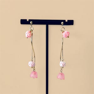 May include: A pair of pink and white dangle earrings with a flower charm. The earrings are made with gold-colored metal and have a delicate, feminine design.