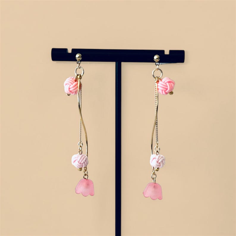 May include: A pair of pink and white dangle earrings with a flower charm. The earrings are made with gold-colored metal and have a delicate, feminine design.