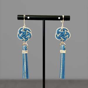 May include: A pair of blue and gold earrings with a tassel. The earrings feature a knotted design and are hanging from a black stand.